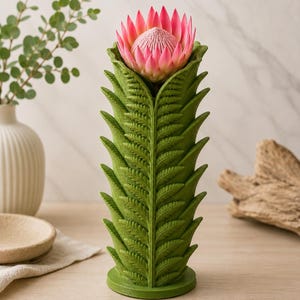 Pu&ograve; includere: Un vaso verde a forma di felce contiene un fiore di protea rosa. Il vaso ha un design dettagliato e strutturato. Il fiore &egrave; di un rosa vibrante con un centro strutturato. Il vaso e il fiore sono su una superficie di legno.