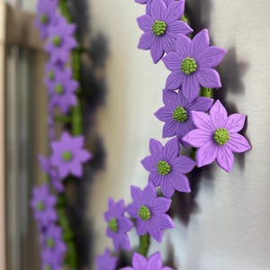 May include: A decorative floral wreath featuring purple flowers with green centers. The flowers are arranged in a circular pattern on a green stem. The wreath is attached to a metallic surface.