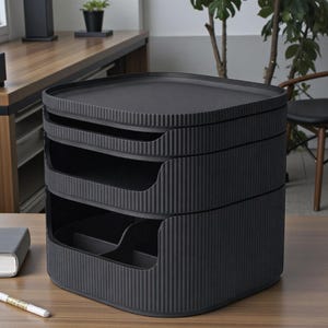 May include: A black, three-tiered desk organizer with a ribbed texture. The top tier is a flat tray, and the lower tiers have open compartments for storage. The organizer is made of a matte black material and sits on a wooden desk.