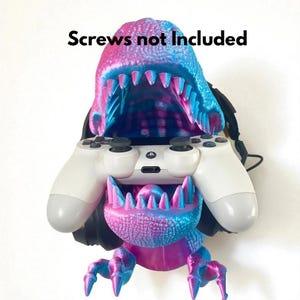 May include: A 3D-printed dinosaur-shaped game controller holder in shades of blue and pink. The holder is designed to cradle a white game controller. The text "Screws not included" is above the dinosaur.