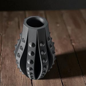 Puede incluir: Un jarr&oacute;n gris oscuro texturizado con un dise&ntilde;o &uacute;nico. El jarr&oacute;n tiene una serie de paneles verticales y curvos con detalles redondeados en relieve y puntas. El jarr&oacute;n est&aacute; sobre un fondo de madera.