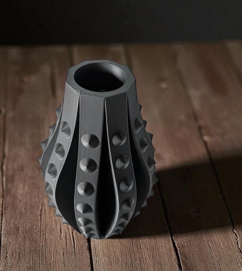 Puede incluir: Un jarr&oacute;n gris oscuro texturizado con un dise&ntilde;o &uacute;nico. El jarr&oacute;n tiene una serie de paneles verticales y curvos con detalles redondeados en relieve y puntas. El jarr&oacute;n est&aacute; sobre un fondo de madera.