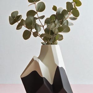 Op de afbeelding: Een moderne, geometrische vaas in zwart, beige en wit, gevormd als een bergketen. De vaas bevat een takje groene eucalyptusbladeren. Een decoratief woonaccessoire.