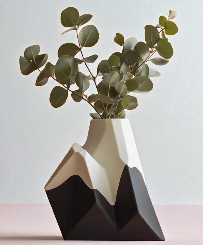 Op de afbeelding: Een moderne, geometrische vaas in zwart, beige en wit, gevormd als een bergketen. De vaas bevat een takje groene eucalyptusbladeren. Een decoratief woonaccessoire.