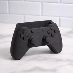 May include: A black desk tidy in the shape of a video game controller. The controller design includes a directional pad, buttons, and analog sticks. The organiser is designed to hold pens, pencils, or other small office supplies. The item is made of a textured material.