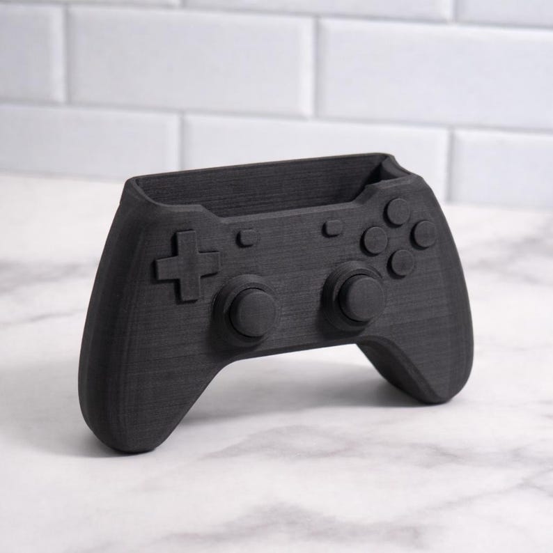 May include: A black desk tidy in the shape of a video game controller. The controller design includes a directional pad, buttons, and analog sticks. The organiser is designed to hold pens, pencils, or other small office supplies. The item is made of a textured material.