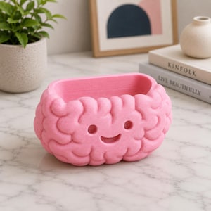 May include: A pink, brain-shaped planter with a smiling face. The planter is made of a textured material and has a rectangular opening. The design is whimsical and suitable for small plants or decorative items.