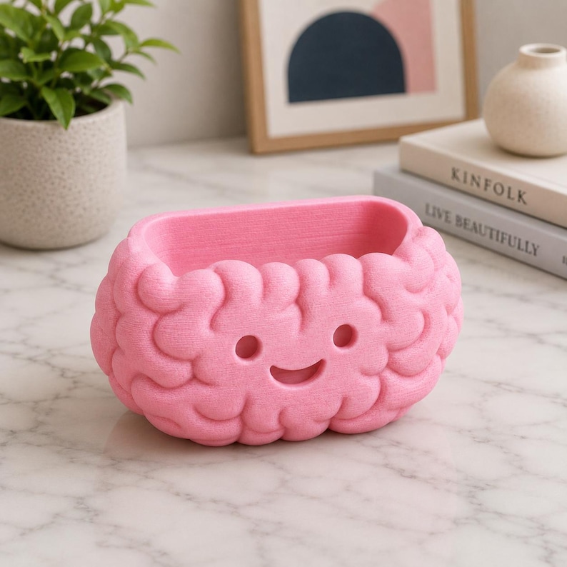 May include: A pink, brain-shaped planter with a smiling face. The planter is made of a textured material and has a rectangular opening. The design is whimsical and suitable for small plants or decorative items.