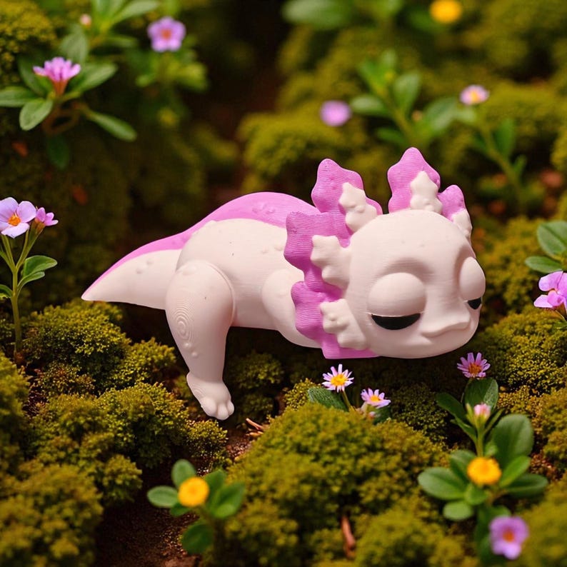 May include: A white and pink axolotl figurine resting on a bed of green moss and small flowers. The axolotl has a detailed design with pink gills and a relaxed expression. The scene is set outdoors, with a natural, whimsical aesthetic.