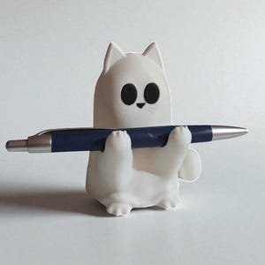 May include: A white cat-shaped pen holder with large black eyes and a small black nose. The holder is holding a dark blue pen with a silver tip. The pen holder is on a white background.