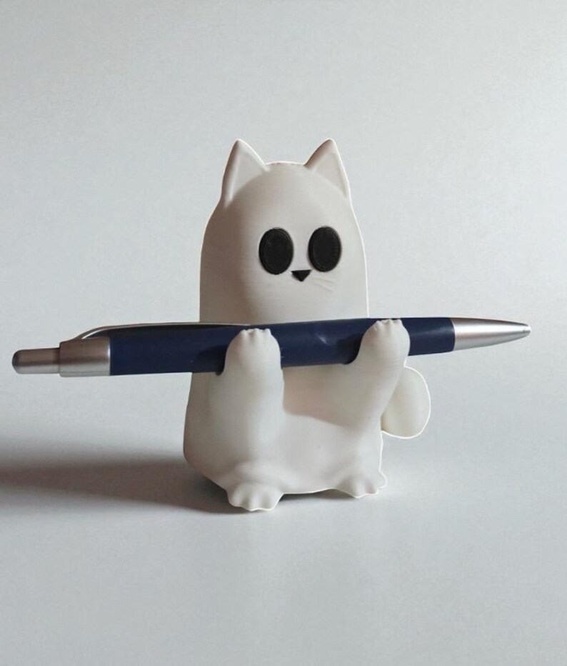 May include: A white cat-shaped pen holder with large black eyes and a small black nose. The holder is holding a dark blue pen with a silver tip. The pen holder is on a white background.