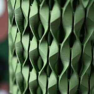 May include: Close-up of a green, geometric, 3D-printed object with a repeating pattern of twisted, diamond-shaped cells. The object has a textured surface and casts shadows, highlighting its intricate design. The overall theme is modern design and decor.