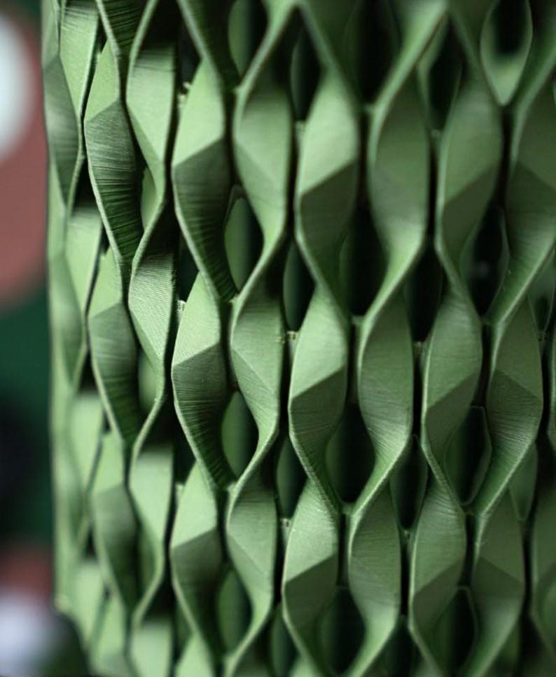 May include: Close-up of a green, geometric, 3D-printed object with a repeating pattern of twisted, diamond-shaped cells. The object has a textured surface and casts shadows, highlighting its intricate design. The overall theme is modern design and decor.