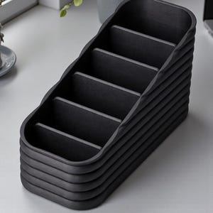 May include: A black desk organizer with multiple compartments. The organizer has a stepped design with a series of horizontal dividers, creating individual storage spaces. The exterior has a textured, ribbed appearance, and the overall shape is rectangular.
