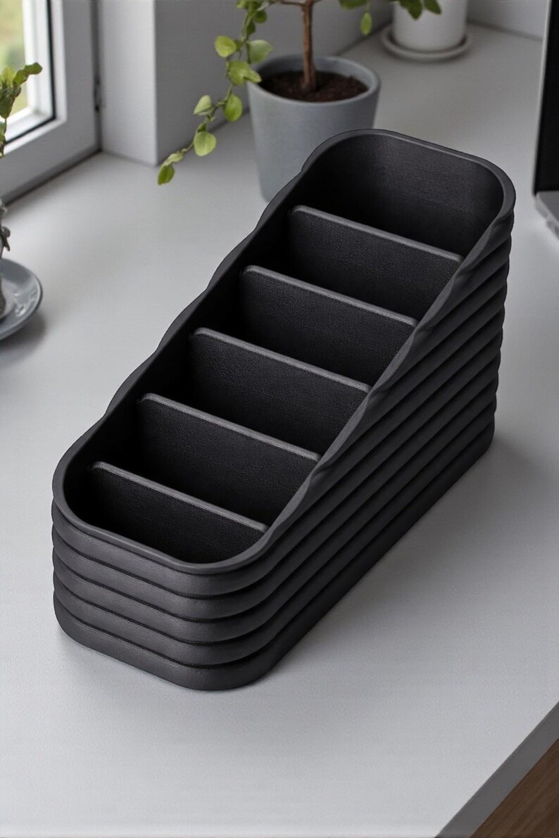 May include: A black desk organizer with multiple compartments. The organizer has a stepped design with a series of horizontal dividers, creating individual storage spaces. The exterior has a textured, ribbed appearance, and the overall shape is rectangular.