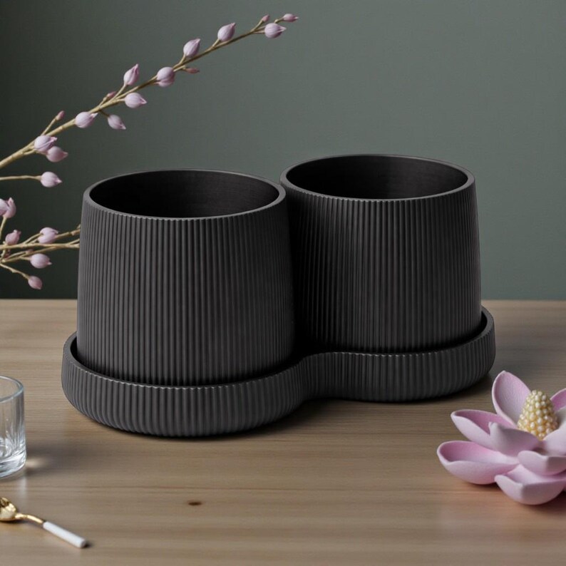 May include: Two dark gray, ribbed ceramic planters with matching saucers. The planters are cylindrical with vertical grooves and sit on a wooden surface. A pink flower and sprig of purple flowers are in the background.