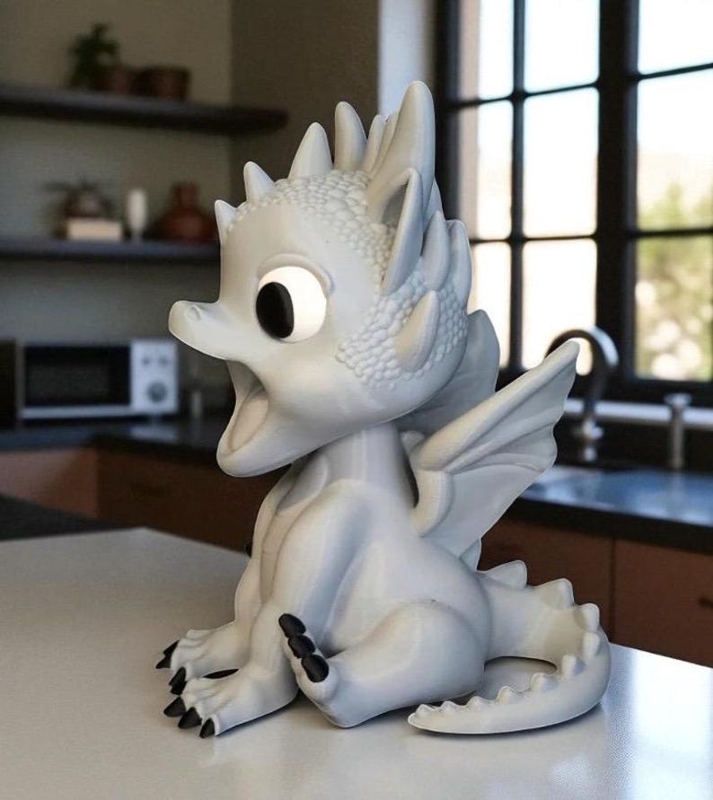 May include: A light gray dragon figurine with large eyes and an open mouth. The dragon has detailed scales, wings, and black claws. It is sitting on a white surface, with a kitchen and window in the background.