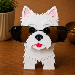 May include: A white dog-shaped eyeglass holder with a black nose, pink ears, and a red tongue. The holder is wearing black sunglasses with a Ray-Ban logo. The holder is made of a textured material and sits on a wooden surface.