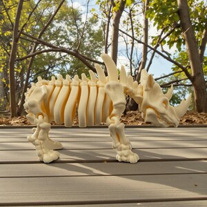 May include: A cream-colored rhinoceros skeleton model. The model is detailed, showing the rib cage, spine, and skull with horns. It stands on four legs, with the background of the image showing trees and a wooden deck.