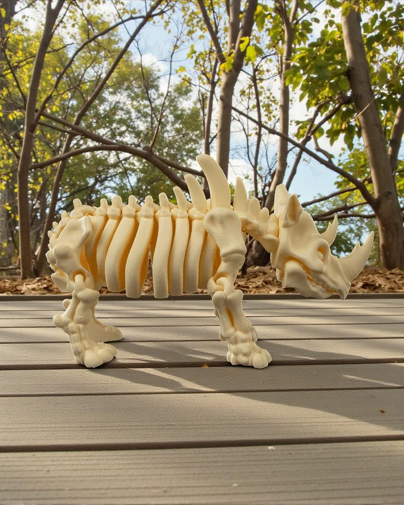 May include: A cream-colored rhinoceros skeleton model. The model is detailed, showing the rib cage, spine, and skull with horns. It stands on four legs, with the background of the image showing trees and a wooden deck.