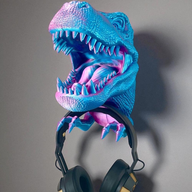 May include: A blue and pink T-Rex head wall mount holding a pair of black and gold headphones. The dinosaur head is open-mouthed, with sharp teeth and a textured surface. The headphones are suspended from the dinosaur's jaw.