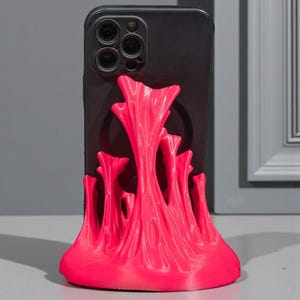 May include: A black smartphone is propped up by a vibrant pink phone stand. The stand has a unique, abstract design with a textured, flowing appearance. The stand is made of a bright, eye-catching pink material.