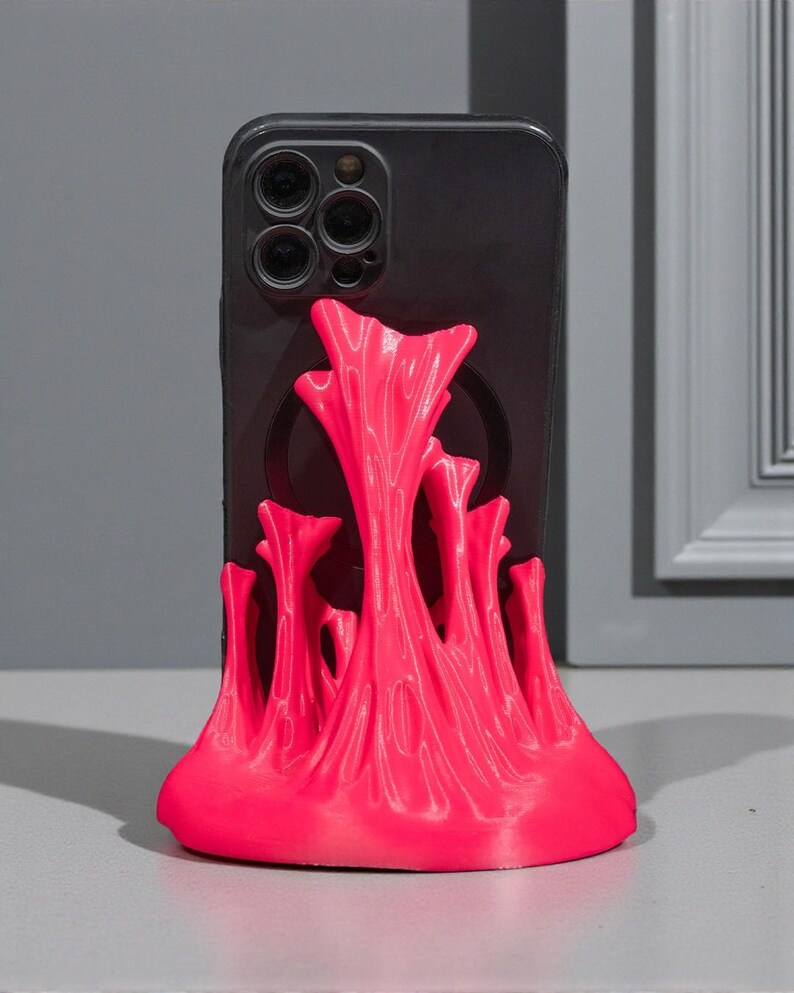 May include: A black smartphone is propped up by a vibrant pink phone stand. The stand has a unique, abstract design with a textured, flowing appearance. The stand is made of a bright, eye-catching pink material.
