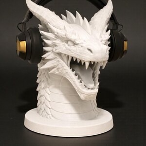 May include: A white dragon head sculpture with an open mouth and sharp teeth, designed to hold headphones. The dragon has detailed scales and horns. Black headphones with gold accents rest on the dragon's head. The sculpture sits on a round base.