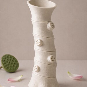 May include: A cream ceramic vase with a textured, bamboo-style design. The vase has four circular, pod-like accents with small holes. Pink and white flower petals and a green seed pod are scattered around the base of the vase.