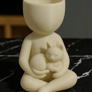 May include: A cream-coloured figurine of a person holding a sleeping cat. The figure has a large head with an open top, possibly for use as a planter or container. The cat is curled up in the figure's lap, creating a serene and whimsical design.