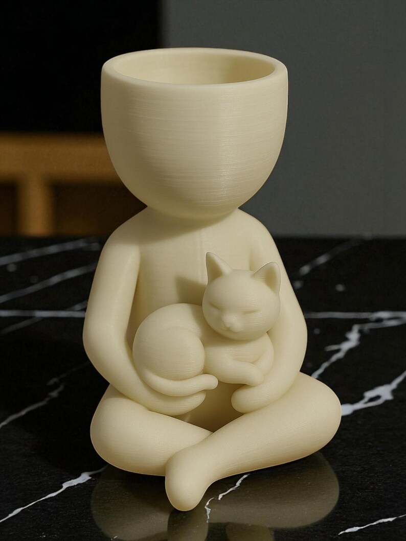 May include: A cream-coloured figurine of a person holding a sleeping cat. The figure has a large head with an open top, possibly for use as a planter or container. The cat is curled up in the figure's lap, creating a serene and whimsical design.