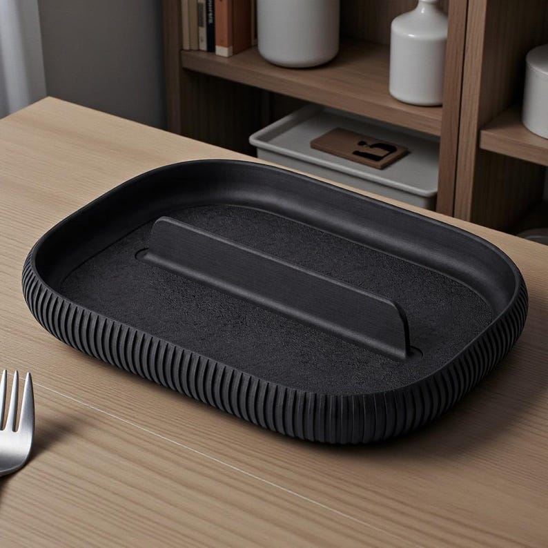 May include: A black rectangular tray with a raised center divider. The tray has a textured surface and a ribbed edge. The tray is sitting on a light brown wooden surface. A silver fork is in the lower left corner.