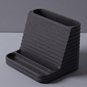 May include: A dark gray desk organizer with a textured, ridged design. The organizer features a sloped back with a rectangular opening and a small tray at the base. Ideal for office or home organization.