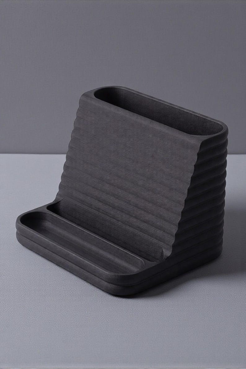 May include: A dark gray desk organizer with a textured, ridged design. The organizer features a sloped back with a rectangular opening and a small tray at the base. Ideal for office or home organization.