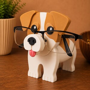 May include: A dog-shaped eyeglass holder in white, tan, and black. The dog has black framed glasses on its nose and a pink tongue sticking out. The holder is on a wooden surface.