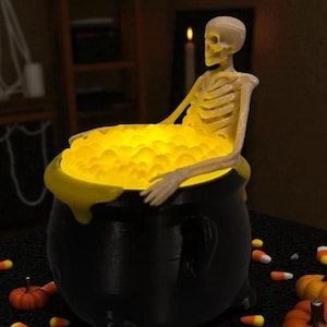 May include: A black cauldron with a yellow glowing liquid, and a skeleton figure sitting inside. The cauldron has a yellow rim and is surrounded by candy corn and miniature pumpkins, creating a Halloween theme.