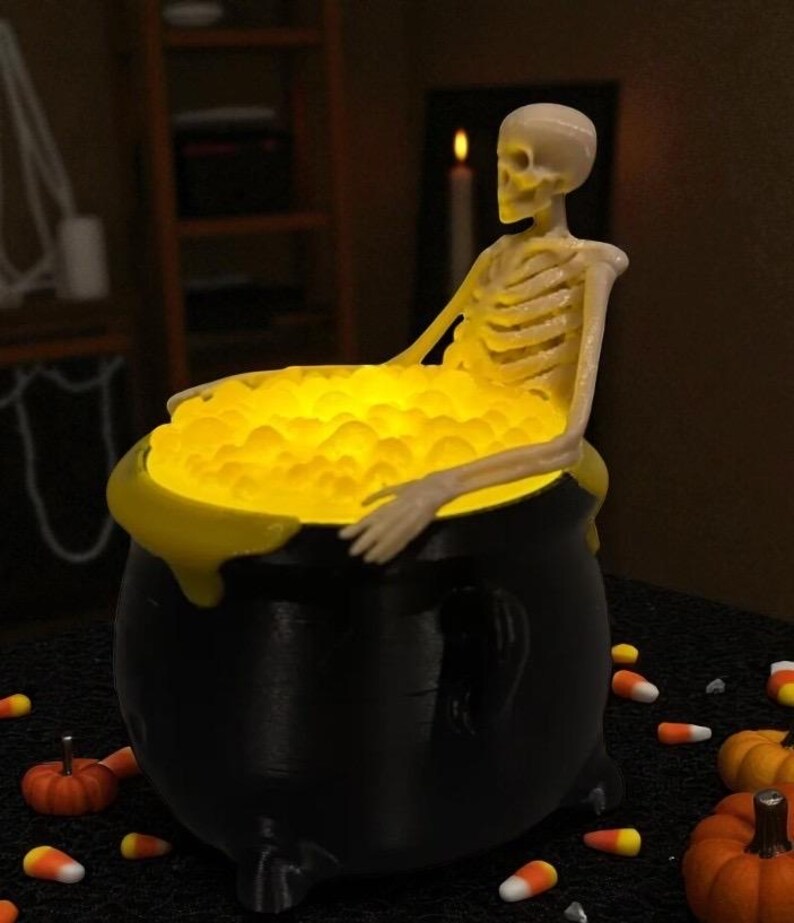 May include: A black cauldron with a yellow glowing liquid, and a skeleton figure sitting inside. The cauldron has a yellow rim and is surrounded by candy corn and miniature pumpkins, creating a Halloween theme.