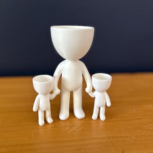 May include: A white, 3D-printed family figurine set. The set includes a large figure holding the hands of two smaller figures. The figures have a minimalist design with a rounded head and body, and are suitable for use as small planters.