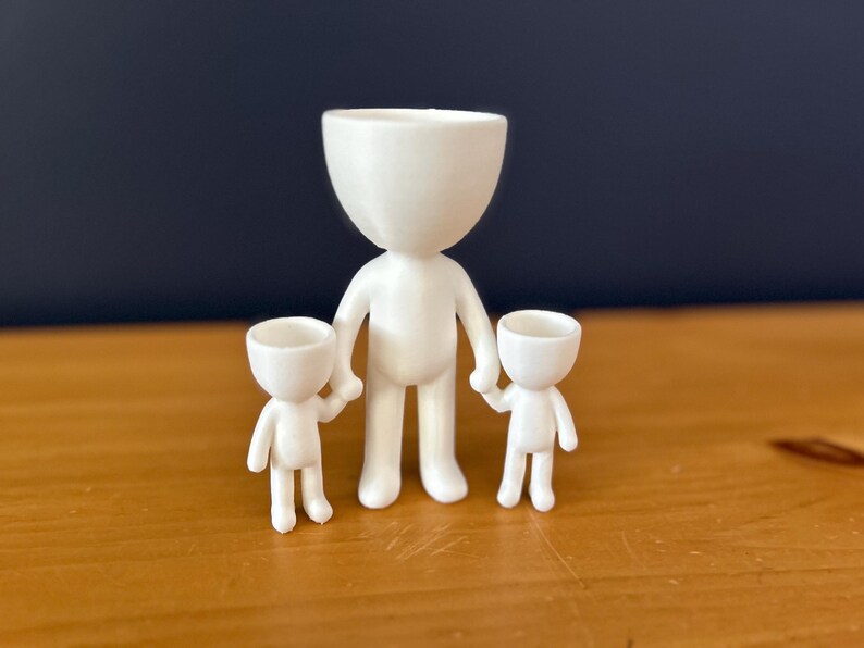 May include: A white, 3D-printed family figurine set. The set includes a large figure holding the hands of two smaller figures. The figures have a minimalist design with a rounded head and body, and are suitable for use as small planters.