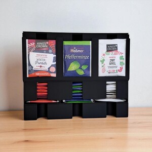 May include: A black tea organizer with three compartments, each holding different tea varieties. The tea packets are in colorful packaging, including "Winter Punch," "Pfefferminze," and "Zimt Apfel." The organizer sits on a wooden surface.