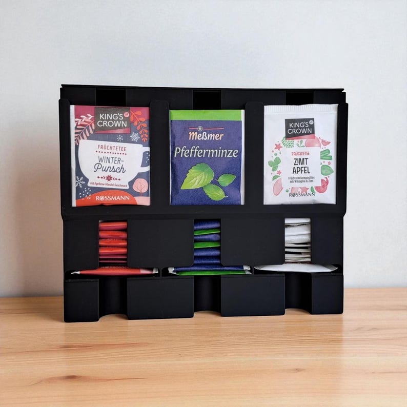 May include: A black tea organizer with three compartments, each holding different tea varieties. The tea packets are in colorful packaging, including "Winter Punch," "Pfefferminze," and "Zimt Apfel." The organizer sits on a wooden surface.