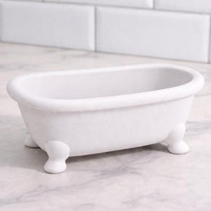 May include: A small, white, claw-foot bathtub. The miniature tub is oval-shaped with rounded edges and sits on four small, rounded feet. The background features a white tile wall and a marble countertop.