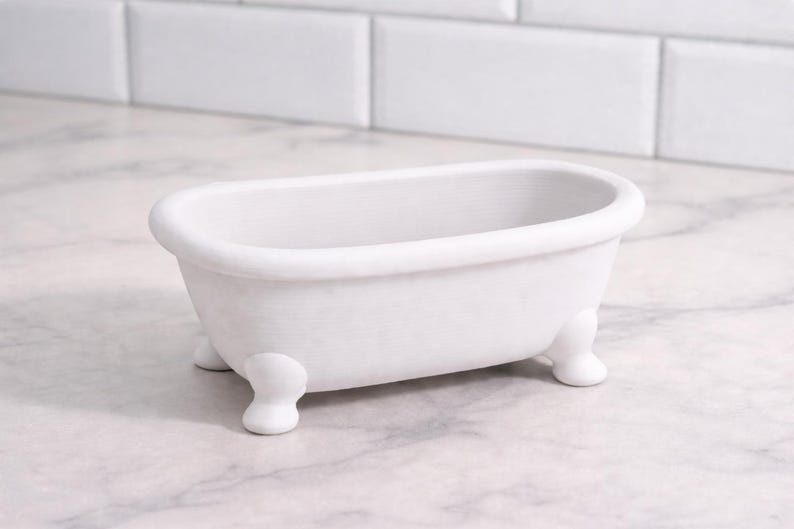 May include: A small, white, claw-foot bathtub. The miniature tub is oval-shaped with rounded edges and sits on four small, rounded feet. The background features a white tile wall and a marble countertop.