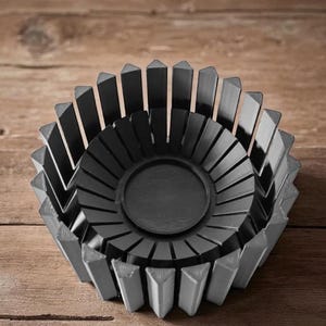 Op de afbeelding: Een donkergrijze decoratieve kom met een geometrisch ontwerp. De kom heeft een cirkelvormig midden en is omgeven door verticale, driehoekige stukken. De kom is gemaakt van een massief materiaal en staat op een houten oppervlak.