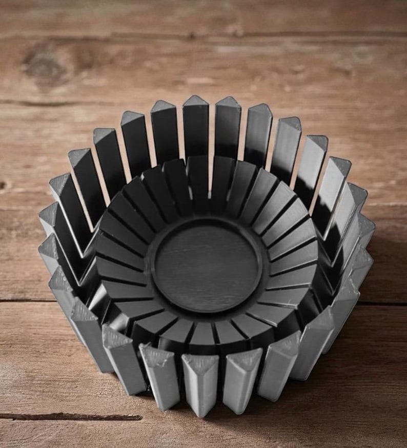 Op de afbeelding: Een donkergrijze decoratieve kom met een geometrisch ontwerp. De kom heeft een cirkelvormig midden en is omgeven door verticale, driehoekige stukken. De kom is gemaakt van een massief materiaal en staat op een houten oppervlak.