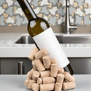 Puede incluir: Una botella de vino verde con una etiqueta blanca en blanco descansa sobre una pila de corchos. La botella est&aacute; en &aacute;ngulo, lo que sugiere que se est&aacute; vertiendo. Los corchos son de color marr&oacute;n claro y cil&iacute;ndricos. El fondo presenta un fregadero y un grifo.