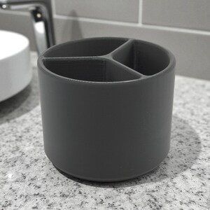 Puede incluir: Un portacepillos cil&iacute;ndrico gris oscuro con tres compartimentos internos. El soporte est&aacute; hecho de un material s&oacute;lido y se encuentra sobre una encimera moteada. El dise&ntilde;o es simple y moderno, adecuado para la organizaci&oacute;n del ba&ntilde;o.
