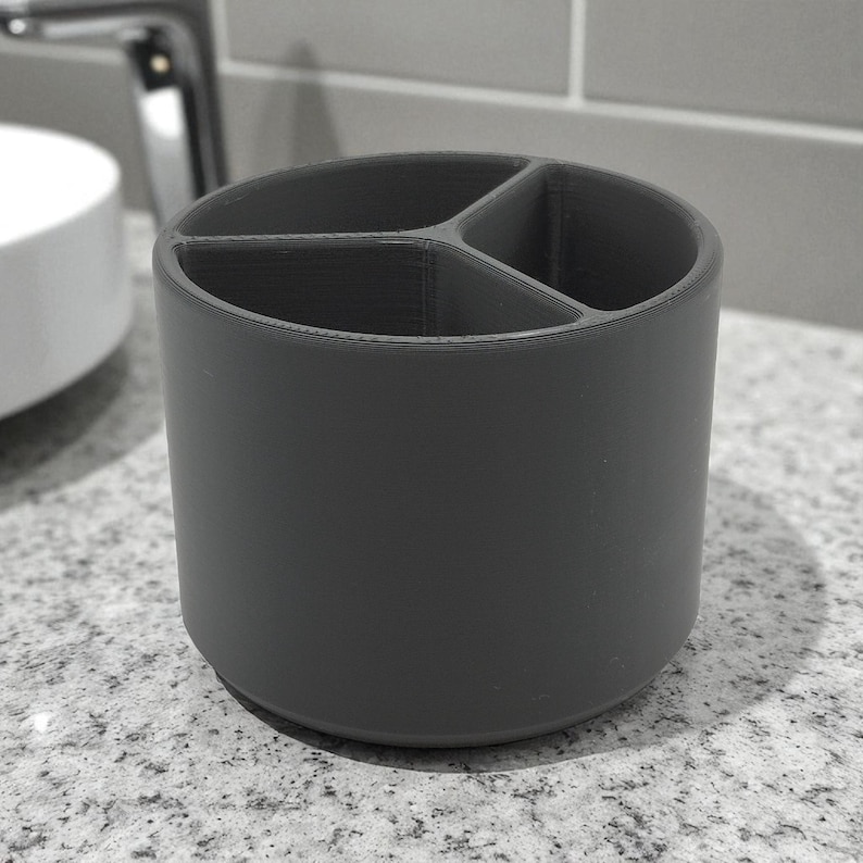 Puede incluir: Un portacepillos cil&iacute;ndrico gris oscuro con tres compartimentos internos. El soporte est&aacute; hecho de un material s&oacute;lido y se encuentra sobre una encimera moteada. El dise&ntilde;o es simple y moderno, adecuado para la organizaci&oacute;n del ba&ntilde;o.