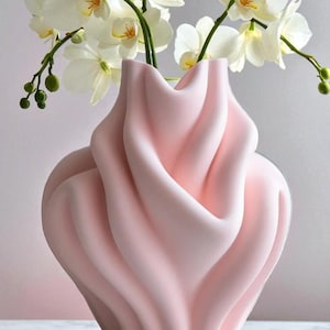 May include: A soft pink, sculptural vase with a flowing, draped design. The vase holds white orchid flowers with yellow centers. The overall aesthetic is modern and elegant, suitable for home decor.
