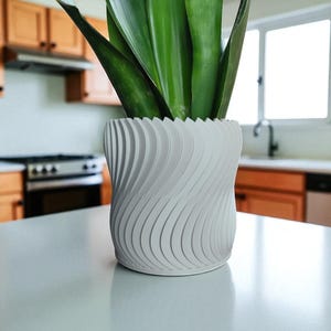May include: A white, modern-style plant pot with a unique, curved design. The pot holds a green plant with long, upright leaves. The pot is sitting on a white countertop in a kitchen setting.